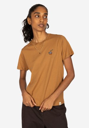 Person mit dunklem lockigem Haar im Pferdeschwanz, trägt ein braunes T-Shirt mit kleinem buntem Schüsseldruck und schwarze Hose, steht vor weißem Hintergrund.