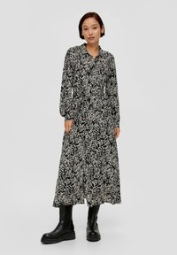 Langes schwarzes Kleid mit abstraktem weißen Blumenmuster, Knopfverschluss und langen Ärmeln; kombiniert mit knielangen schwarzen Stiefeln.