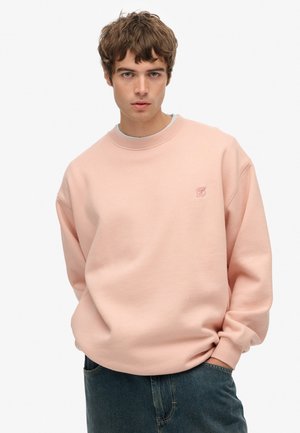 Sweatshirt rose clair avec un col rond, des poignets côtelés et un petit logo brodé sur la poitrine. Le tissu semble doux et texturé.