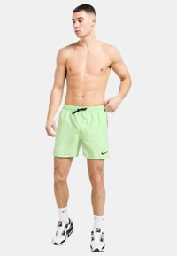 Pantaloni da bagno verde neon con vita regolabile, tasca laterale e logo Nike nero. Indossati con scarpe e calze sportive bianche e nere.