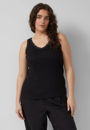 Frau mit langen lockigen Haaren, die ein schwarzes ärmelloses Oberteil mit Spitzenbesatz und schwarze weite Hosen trägt, steht vor einem schlichten grauen Hintergrund.