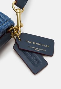 Étiquettes de clés en cuir bleu attachées à un fermoir doré, l'une embossée "THE SOHO FLAP LIMITED RE-EDITION 2006", connectée à un tissu en denim.