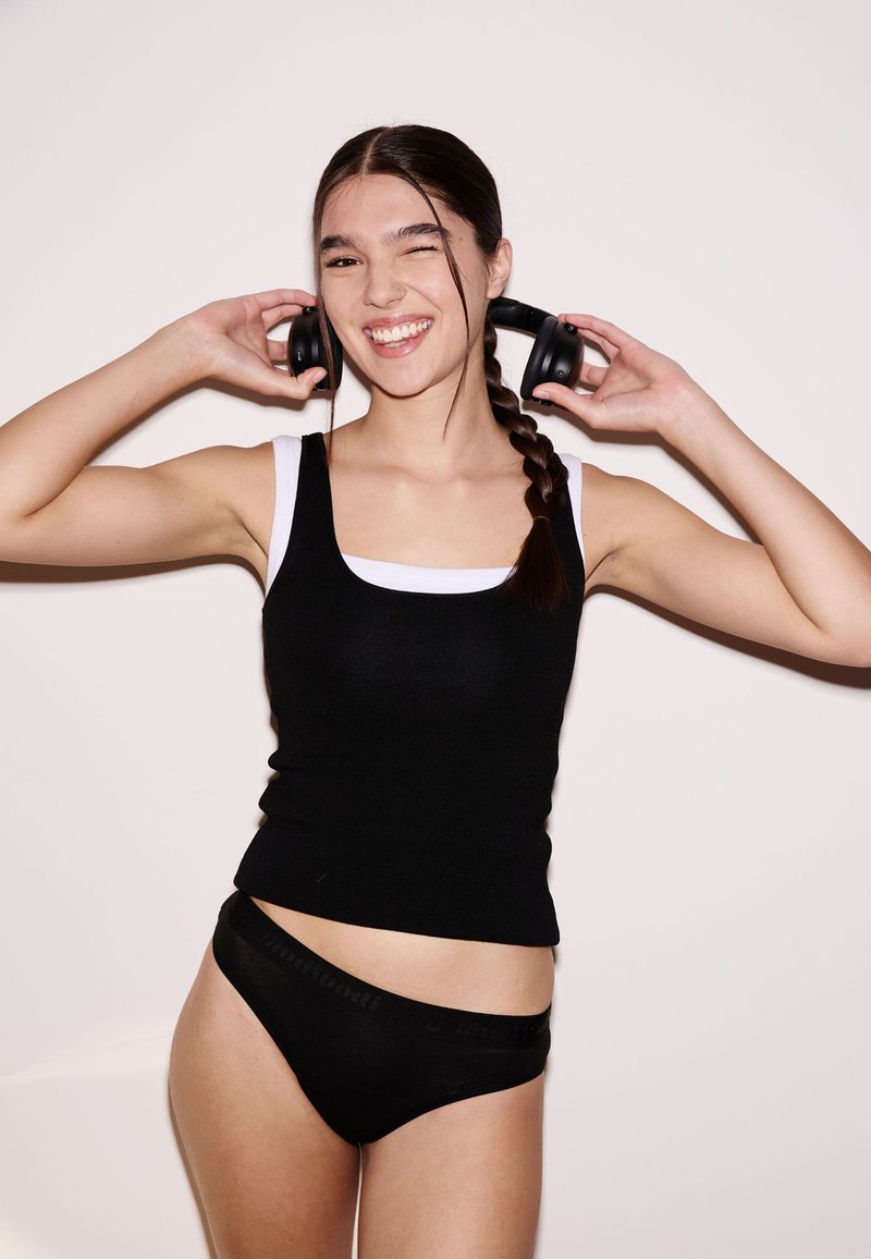 Schwarzes Tanktop mit weißen Trägern, kombiniert mit schwarzer Unterwäsche. Das Model trägt einen Zopf, schwarze Kopfhörer und lächelt vor hellem Hintergrund.