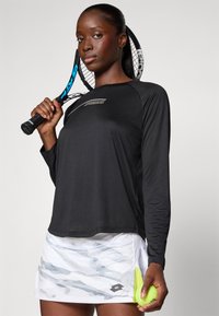 Zwarte sporttop met lange mouwen en een gladde textuur, gecombineerd met een rok met wit camouflagemotief en een neongele accent. Er wordt een tennisracket en bal vastgehouden.