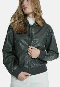 Giacca bomber in pelle nera con polsini e fondo a costine, chiusura con zip e tasche laterali; texture liscia con design minimalista.