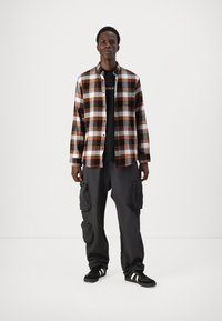 Geruite flanellen overhemd in oranje, zwart en wit, gecombineerd met donkere cargobroek. Model draagt zwarte sneakers met witte strepen.