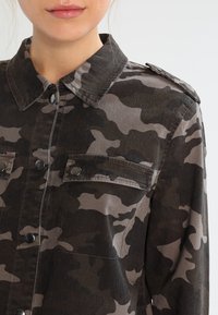 Kvinna som bär en mörkgrå och svart kamouflageklänning med knappar, framfickor med lock och axelklaffar.