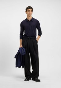 Młody mężczyzna w granatowej koszuli polo z długim rękawem, czarnych szerokich spodniach, czarnych butach, trzymający granatową kurtkę, stojący na tle jasnego gładkiego tła.