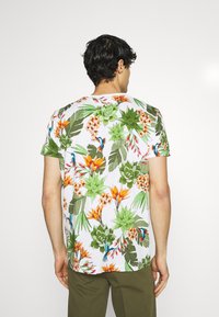 Camiseta de manga corta con un vibrante estampado floral tropical en verde, naranja y azul sobre un fondo blanco. Cuello redondo, corte relajado.