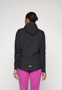 Schwarze wasserdichte Jacke mit Kapuze, ausgestattet mit einer glatten Textur und einem Logo auf dem Rücken, kombiniert mit eng anliegenden pinken Leggings.