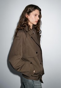 Brown, oversized jacket with a double-breasted design, large buttons, and side pockets, paired with light blue jeans.