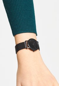 Zwarte polshorloge met een ronde wijzerplaat en een minimalistisch ontwerp. Beschikt over een gevlochten metalen band en een matte afwerking op de wijzerplaat.