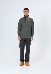 Man staat en draagt een donkergroene trui met knopen en witte trekkoorden, donkere spijkerbroek en tankleurige laarzen, tegen een witte achtergrond.