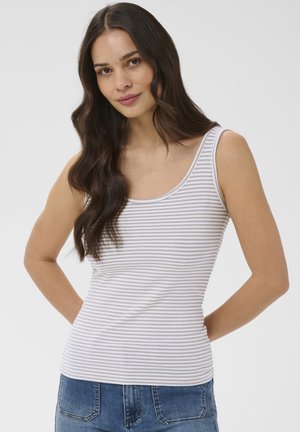 Kvinde med langt mørkt hår iført en hvid og lysegrå stribet ærmeløs top og blå jeans, stående med hænderne bag ryggen.