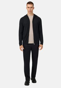 Felpa nera con zip e finitura testurizzata, indossata sopra una maglietta beige chiaro, abbinata a pantaloni neri e sneakers nere.