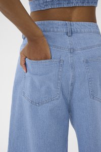 Pantalon en denim bleu clair avec une coupe décontractée, comportant deux poches arrière et une taille. Le tissu a une apparence texturée.
