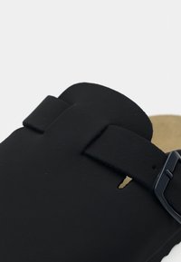 Κοντινό πλάνο λουριού μαύρης δερμάτινης σάνδάλας με ματ μαύρο μεταλλικό αγκράφα και μπεζ πάτο στο φόντο.