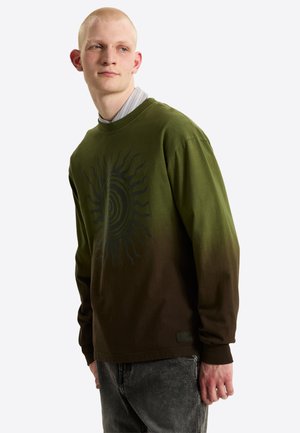 Joven con sudadera de degradado de verde a marrón con diseño de sol, sobre una camisa a rayas, mirando hacia un lado.