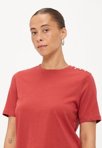 Rode katoenen T-shirt met een ronde hals en korte mouwen, met drie decoratieve knopen op de schouder. Soepele textuur, relaxte fit.