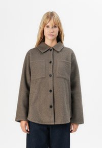 Vrouw in een beige, gestructureerde jasje met knopen en twee borstzakken, met donkerblauwe broek, tegen een effen witte achtergrond.