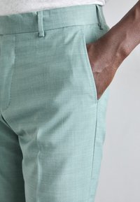 Šviesiai žali kelnės, pagamintos iš tekstūruoto audinio, turinčios šoninį kišenę su matoma ranka, pabrėžiančia jų laisvalaikio dizainą ir pritaikymą.