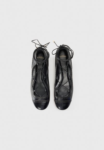 Black leather ballet flats with lace-up design, rounded toe, and decorative perforated detailing along the upper edge. Smooth texture.