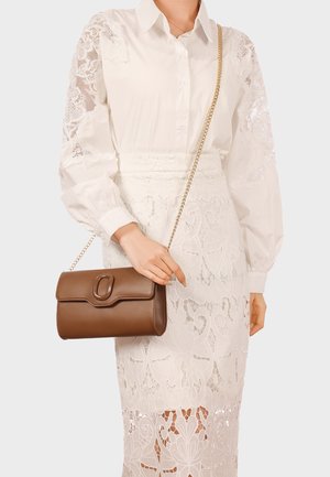 Donna che indossa una blusa bianca con maniche di pizzo e una gonna di pizzo bianca, che tiene una borsa a tracolla in pelle marrone con tracolla a catena dorata.