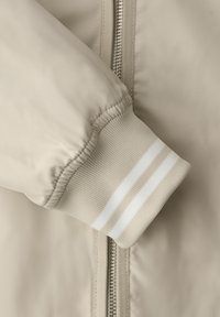 Manga de chaqueta beige con puño elástico acanalado que presenta dos rayas blancas, mostrado sobre una cremallera plateada parcialmente visible en tejido a juego.