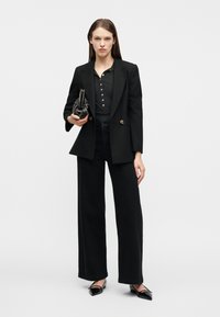 Γυναίκα που φοράει μαύρο σακάκι, μαύρο πουκάμισο με κουμπιά, μαύρο παντελόνι φαρδύ, κρατάει μια μαύρη τσάντα και μαύρα παπούτσια με μυτερό άκρο, στέκεται σε λευκό φόντο.