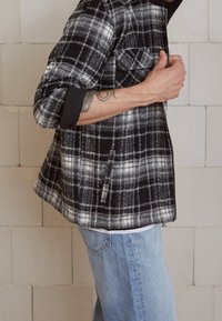 Giacca a scacchi in bianco e nero con orlo arrotondato, due tasche sul petto e polsini neri, abbinata a jeans azzurri chiari.