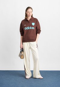 Barna kapucnis felső kék "adidas" logóval és gallérral, krém színű nadrággal, amelynek oldalán piros csíkok találhatók. A modell egy táskát tart. Egyszerű háttér.