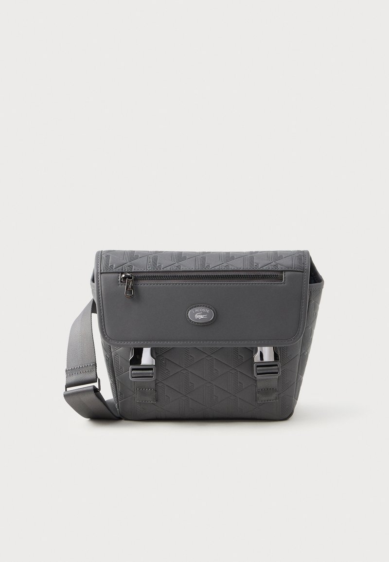 Bolsa crossbody cinza da Lacoste com padrão geométrico, aba frontal com bolso com fecho de zip e fechos de fivela metálica, além de uma alça de ombro ajustável.