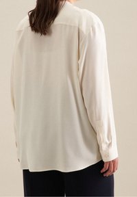 Světle krémová košile s dlouhým rukávem a límečkem, hladká textura a manžety na knoflíky, zobrazená zezadu na neutrálním pozadí.