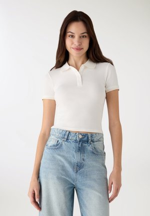 Polo bianca a maniche corte con texture a coste e abbottonatura a tre bottoni, abbinata a jeans azzurri a vita alta.