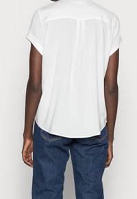 Personne portant un blouse blanche à manches courtes avec des poignets retroussés et un jean en denim bleu, vue de dos sur un fond blanc.