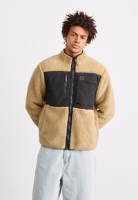 Tarn- und schwarze Fleecejacke mit hohem Kragen, Reißverschluss und einer Brusttasche; weiche Textur mit kontrastierenden Paneelen.