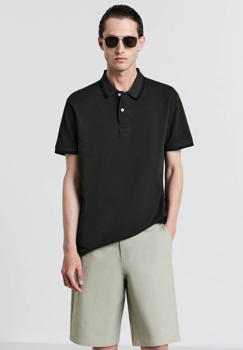 Uomo che indossa una polo nera, pantaloni beige e occhiali da sole, in piedi con una mano in tasca contro uno sfondo neutro.