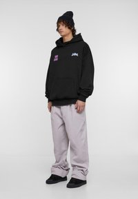 Fekete kapucnis pulóver rózsaszín és kék grafikai szöveggel, laza világosszürke kordbársony nadrággal és fekete cipővel párosítva. A modellen sapka van.