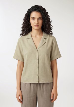 Donna con capelli ricci e scuri che indossa una camicia a maniche corte color beige chiaro con bottoni e pantaloni larghi color taupe, in piedi contro uno sfondo bianco.
