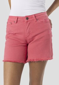 Różowe szorty z dżinsu z postrzępionym wykończeniem, z przetarciami i metalowym zapięciem na guzik. Przednie kieszenie widoczne po obu stronach.