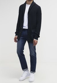 Svart stickad cardigan med sjalkrage, knappframsida och ribbade ärmslut, parat med blå jeans och vita sneakers.