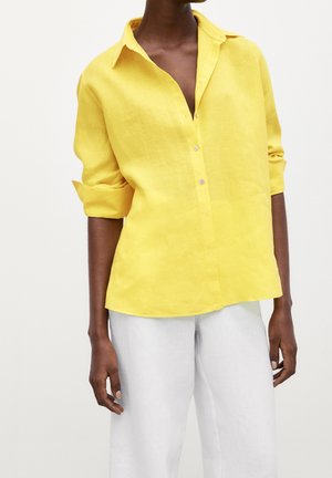 Modèle portant une chemise jaune ample à boutons avec manches retroussées et un pantalon blanc sur un fond clair uni.