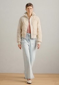 Töltött krémszínű rövid kabát, prémmel díszítve, világoskék, bővített farmernadrággal párosítva. A modellen rózsaszín felső és fehér lapos cipő van.