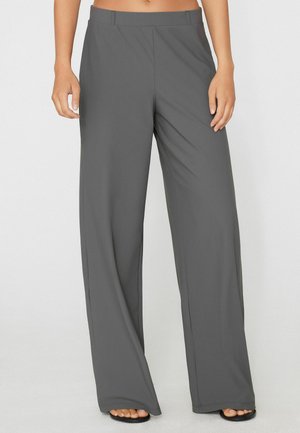 Donna che indossa pantaloni grigi a vita alta e a gamba larga abbinati a sandali neri con punta aperta, in piedi davanti a uno sfondo chiaro e semplice.