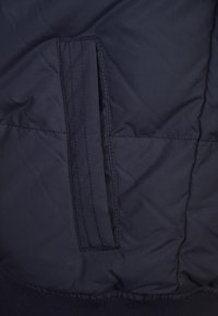 Navy blue puffer jacket fabric with a smooth texture. Features a side pocket with vertical opening and stitched detailing.