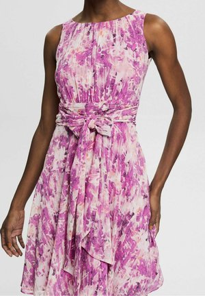 Ærmeløs lyserød og hvid blomsterkjole med plisseret bodice og et bindebælte i taljen på en person med mørk hud mod en ensfarvet baggrund.