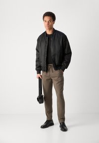 Crna bomber jakna s glatkom teksturom, uparena s bež hlačama i crnim cipelama. Model drži crnu torbu na remenu.