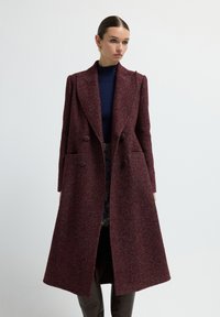 Abrigo burdeos de doble botonadura con patrón de espiga, hecho de tela texturizada. Presenta solapas grandes y bolsillos laterales, con una silueta larga.