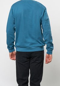 Tyrkysová mikina se zaobleným výstřihem, dlouhými rukávy a žebrovanými manžetami. Kombinováno s černými kalhotami s kapsami na zadní straně. Hladká textura.