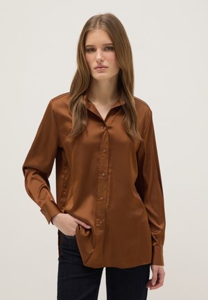 Joven mujer con cabello liso castaño claro que lleva una camisa marrón de satén con mangas largas y botones, y pantalones oscuros, con una mano en el bolsillo.